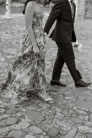 A black and white photo of a couple walking on cobblestones, showing a floral dress and suit.