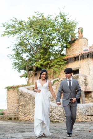A couple in wedding attire walks hand in hand along a rustic stone wall path in a romantic outdoor setting.
