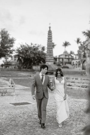 A couple in wedding attire walks together on a grassy path with a pagoda visible in the background.