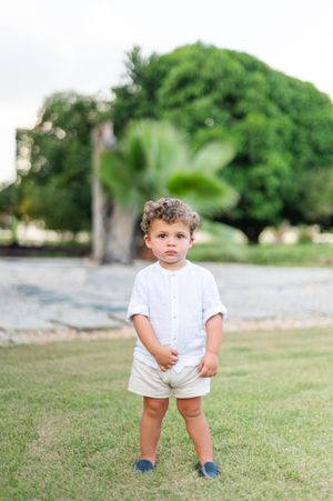 Small toddler wearing white outfit and blue shoes stands on grassy lawn in outdoor setting.