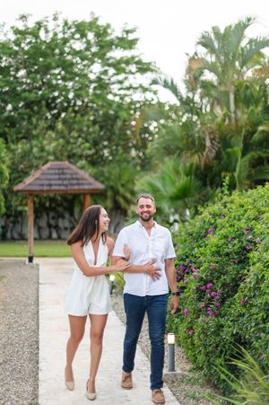 A couple in white outfits walks hand in hand along a tropical garden path surrounded by lush greenery and flowers.