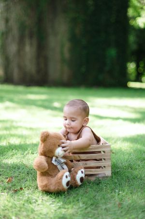 A baby lies on grass near a wooden crate with a teddy bear in a series of progressive shots.