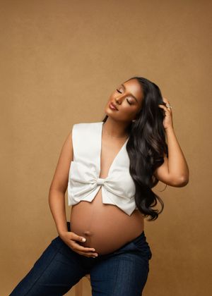 Maternity portraits showing expectant mother in white crop top and jeans against warm brown backdrop.