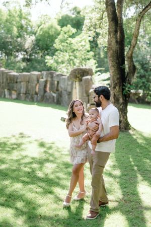 A family enjoys a playful moment together in a sunlit park with trees casting dappled shadows on the grass.