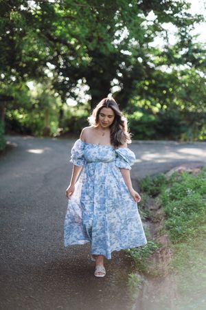 Woman in light blue floral off-shoulder dress with puff sleeves walks along a tree-lined path on a sunny day.