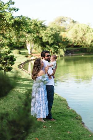 A family stands together on a grassy bank beside a tranquil lake surrounded by lush green trees on a sunny day.