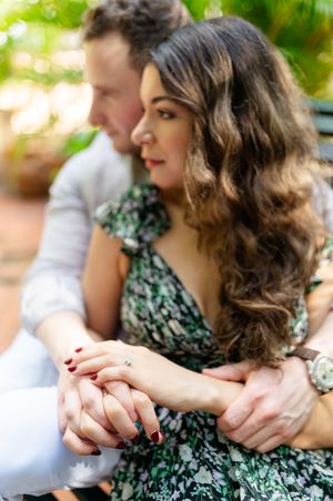 A couple holds hands tenderly while sitting together, with the woman wearing a green floral dress and styled wavy hair.