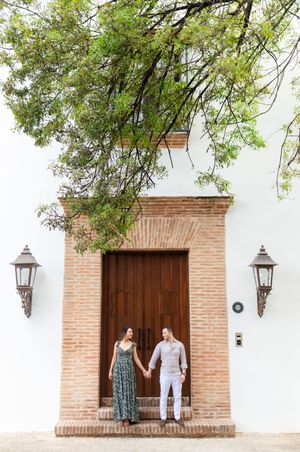 A couple stands in front of an elegant Spanish colonial doorway with brick trim and decorative lanterns on a white facade.