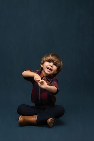 A young child in a burgundy shirt and dark pants sits playfully on a dark studio backdrop.