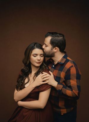 A romantic couple in a plaid shirt and burgundy dress embrace against a dark brown studio backdrop.