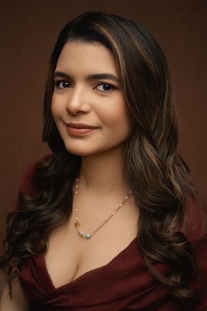 Professional portrait in burgundy top with wavy brown hair and delicate necklace against dark background.