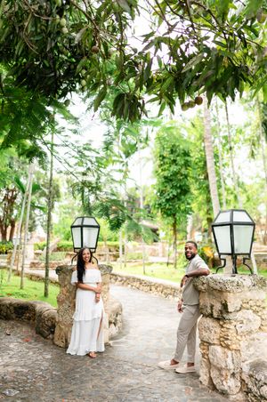 Two people stand on a stone-lined garden path surrounded by lush greenery and decorative lanterns in a romantic setting.