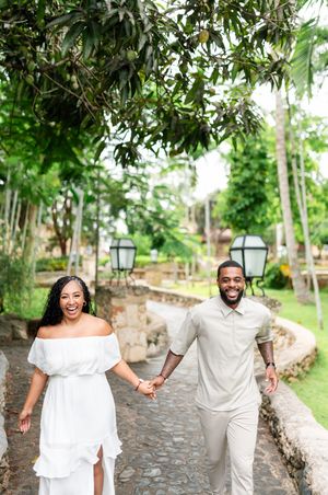 A joyful couple wearing white outfits walks hand in hand along a cobblestone path surrounded by lush greenery.