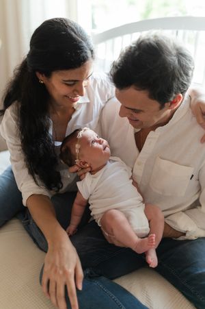 Parents share a tender moment with their newborn baby while sitting together on a couch by a window.