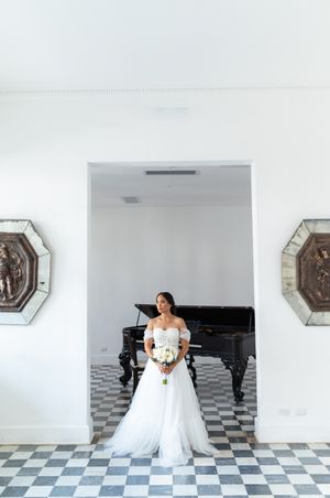 A bride in a flowing white gown poses elegantly on a black and white checkered floor in a bright indoor space.