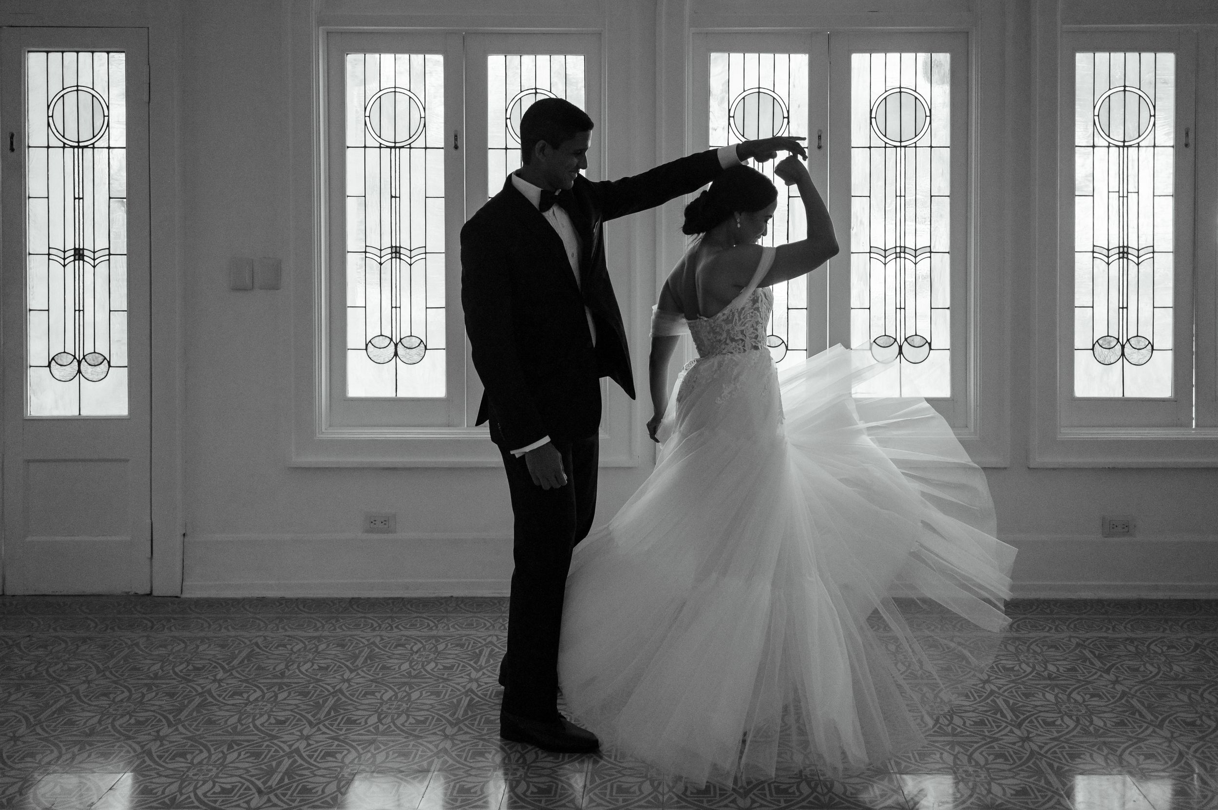 A romantic black and white dance moment between newlyweds in a bright room with ornate windows.