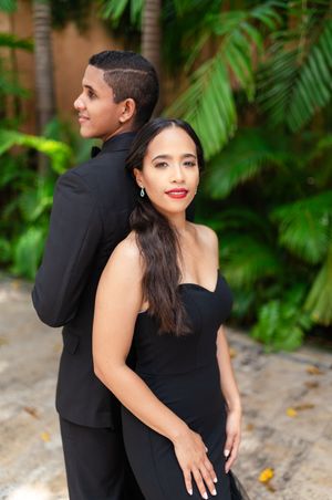 A couple in formal black attire poses together against a lush tropical garden backdrop.