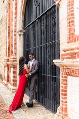 Romantic couple in red dress and gray suit embraces against rustic brick wall with black iron gates.