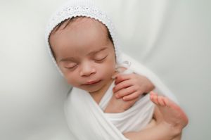 Sleeping newborn baby wearing white bonnet and wrap rests peacefully on soft white fabric.