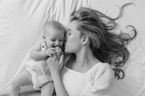 A tender black and white portrait of a mother and baby lying together on white fabric with flowing hair.