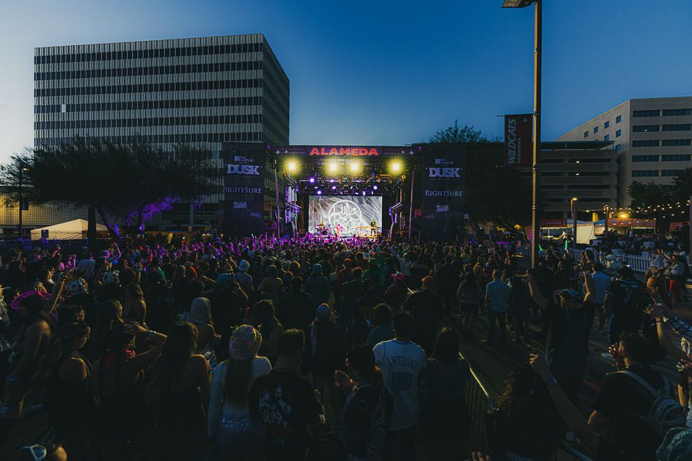 Large crowd gathered at an outdoor concert venue during sunset with city buildings in the background.