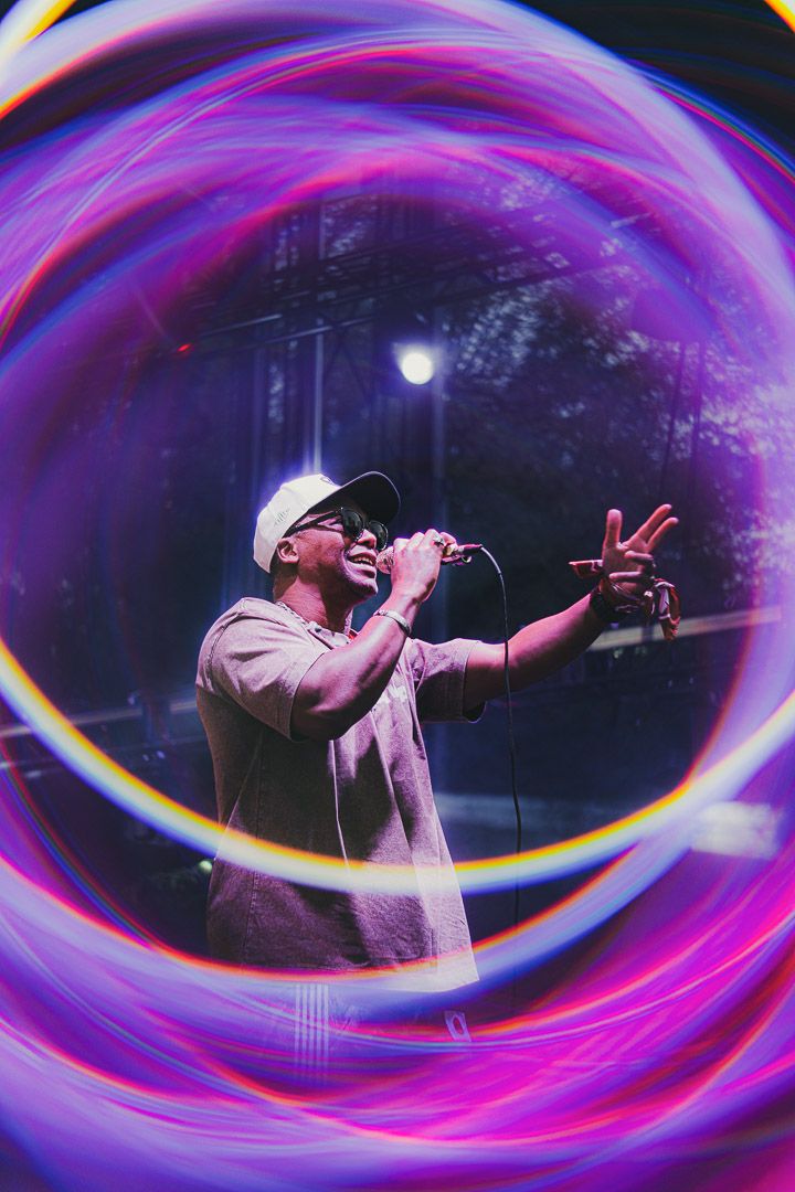 Concert performer in white cap and shirt silhouetted against dark stage with vibrant purple and pink light swirls effect.