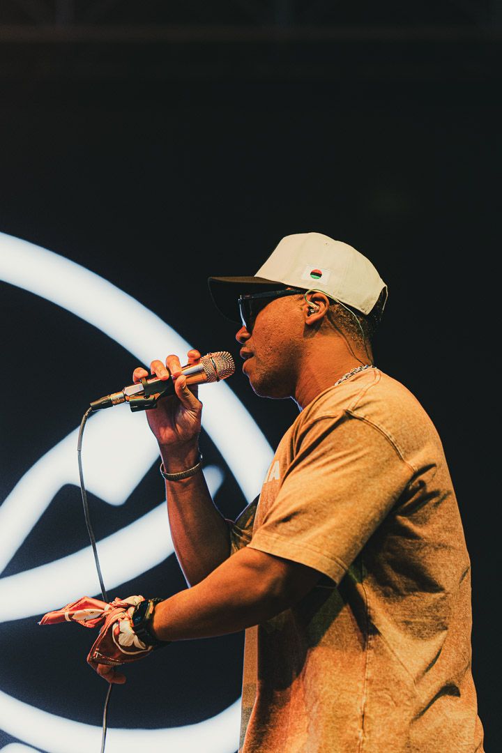 Performer in tan shirt and white cap singing into microphone against dark background with curved light design.