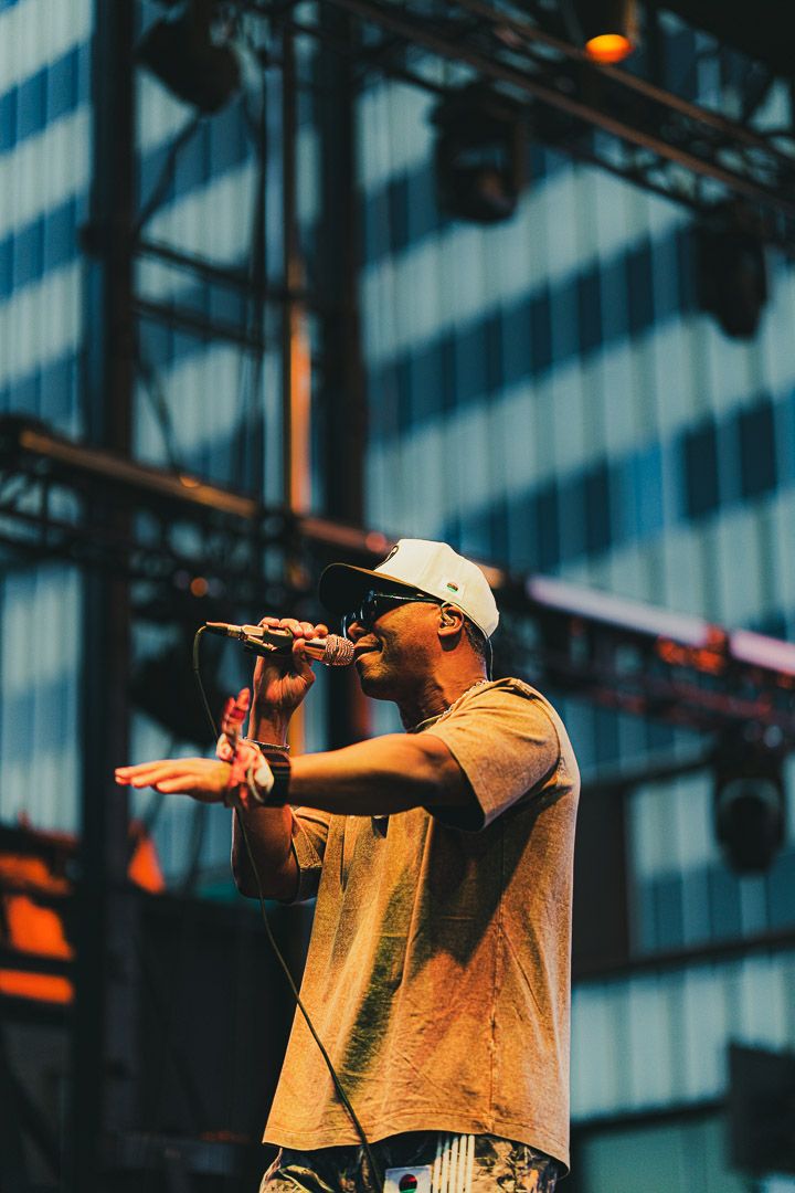 Concert performer in beige shirt and baseball cap gesturing while singing on outdoor stage with city backdrop.