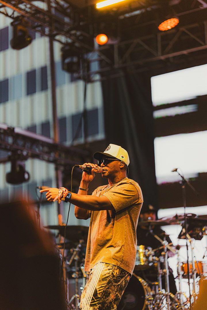 Artist performing on outdoor festival stage wearing tan shirt and white cap with lighting rigs visible above.