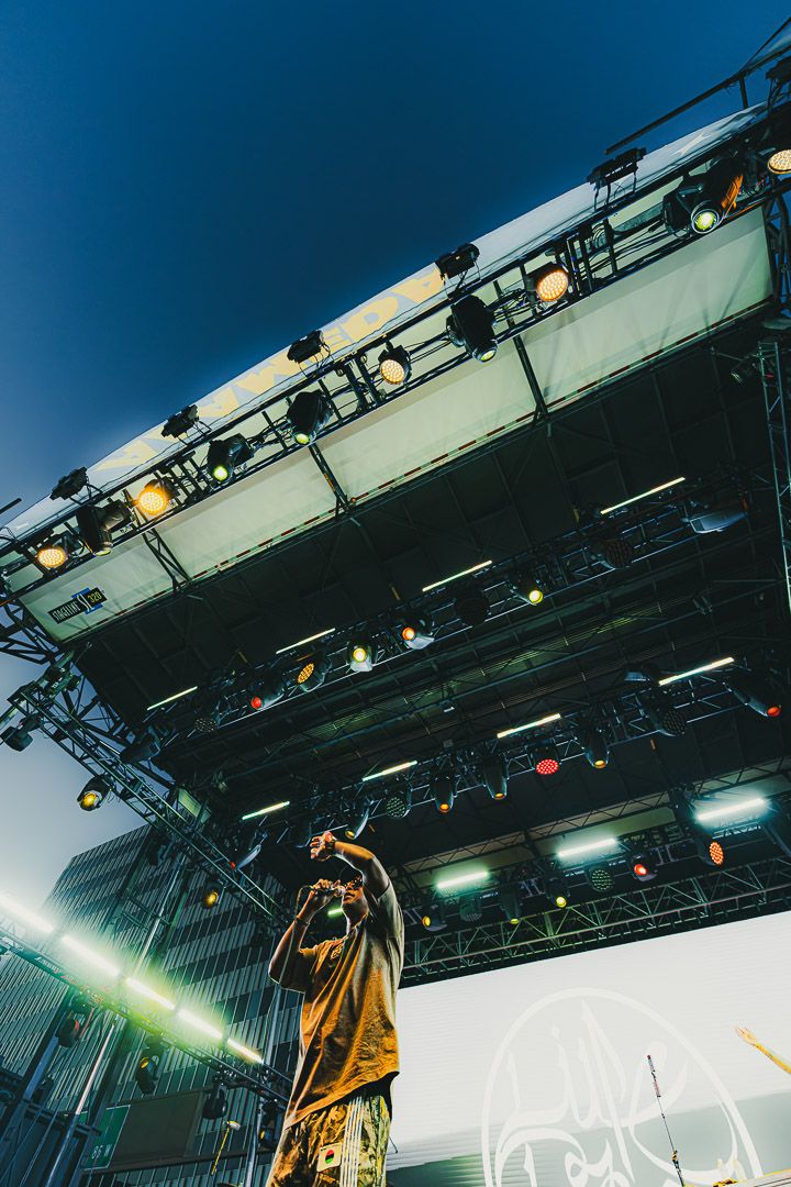 Low-angle concert stage view showing performer in yellow hoodie with dramatic lighting and overhead rigging equipment.