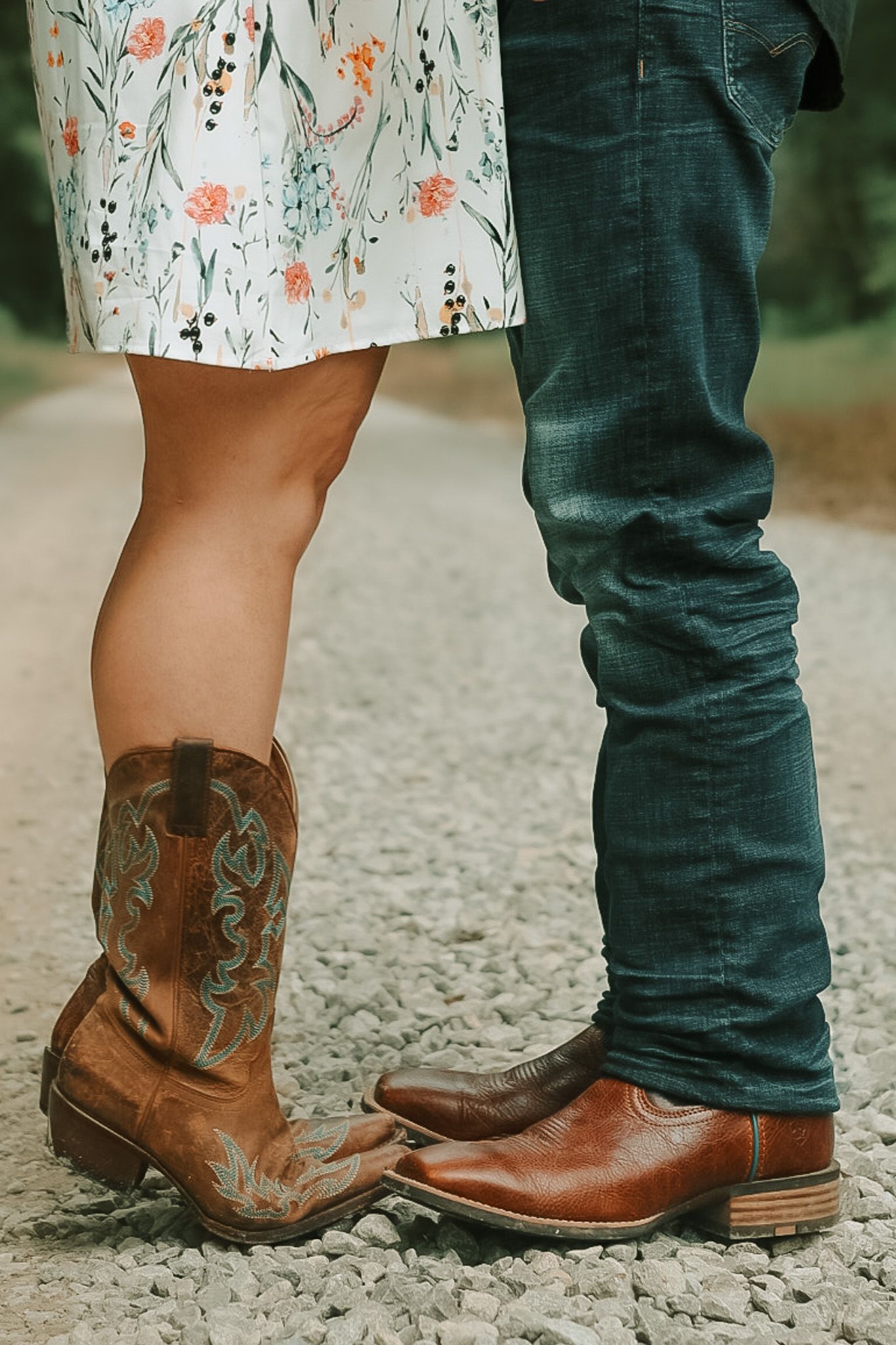 A couple's feet in cowboy boots, one wearing a floral dress and the other in dark jeans, standing close together.