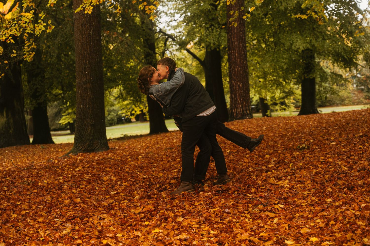 Queer couple walking through vibrant orange autumn leaves scattered on ground beneath tall trees in a park setting.