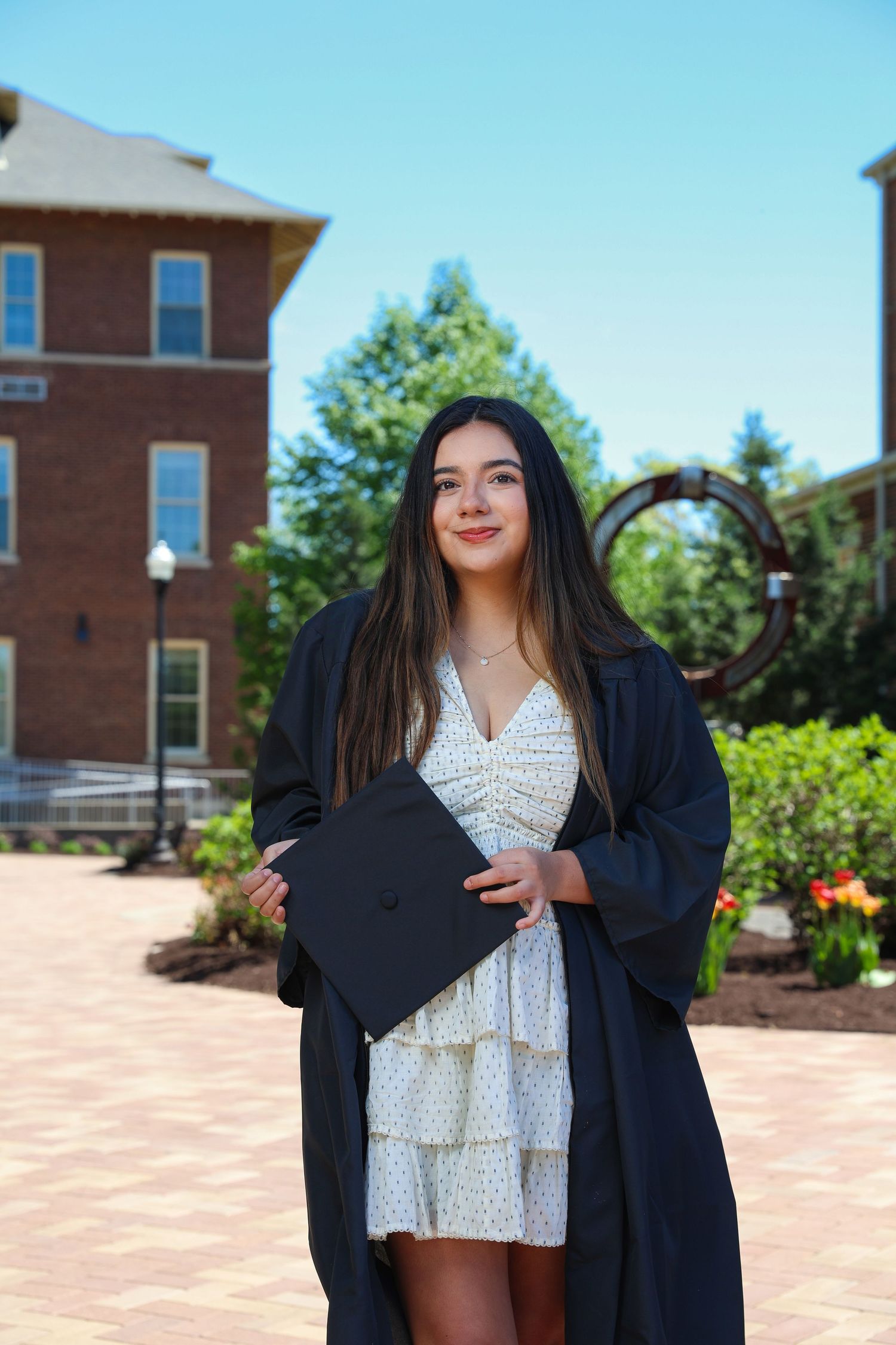 Graduate in black cap and gown poses with white polka dot dress against brick building backdrop on sunny day.