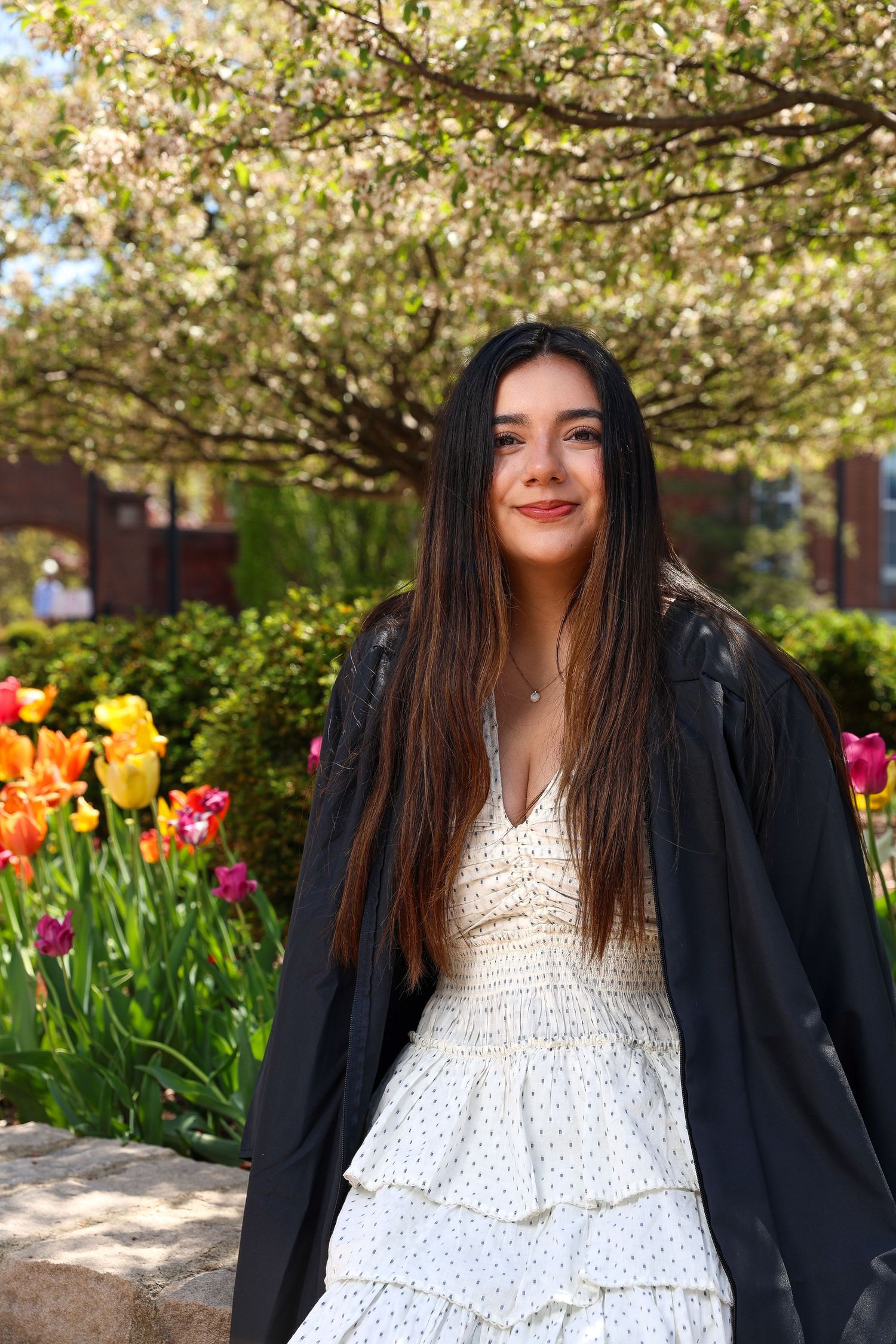 A person in a white polka dot dress and black jacket poses near blooming cherry blossoms and colorful tulips in a garden.
