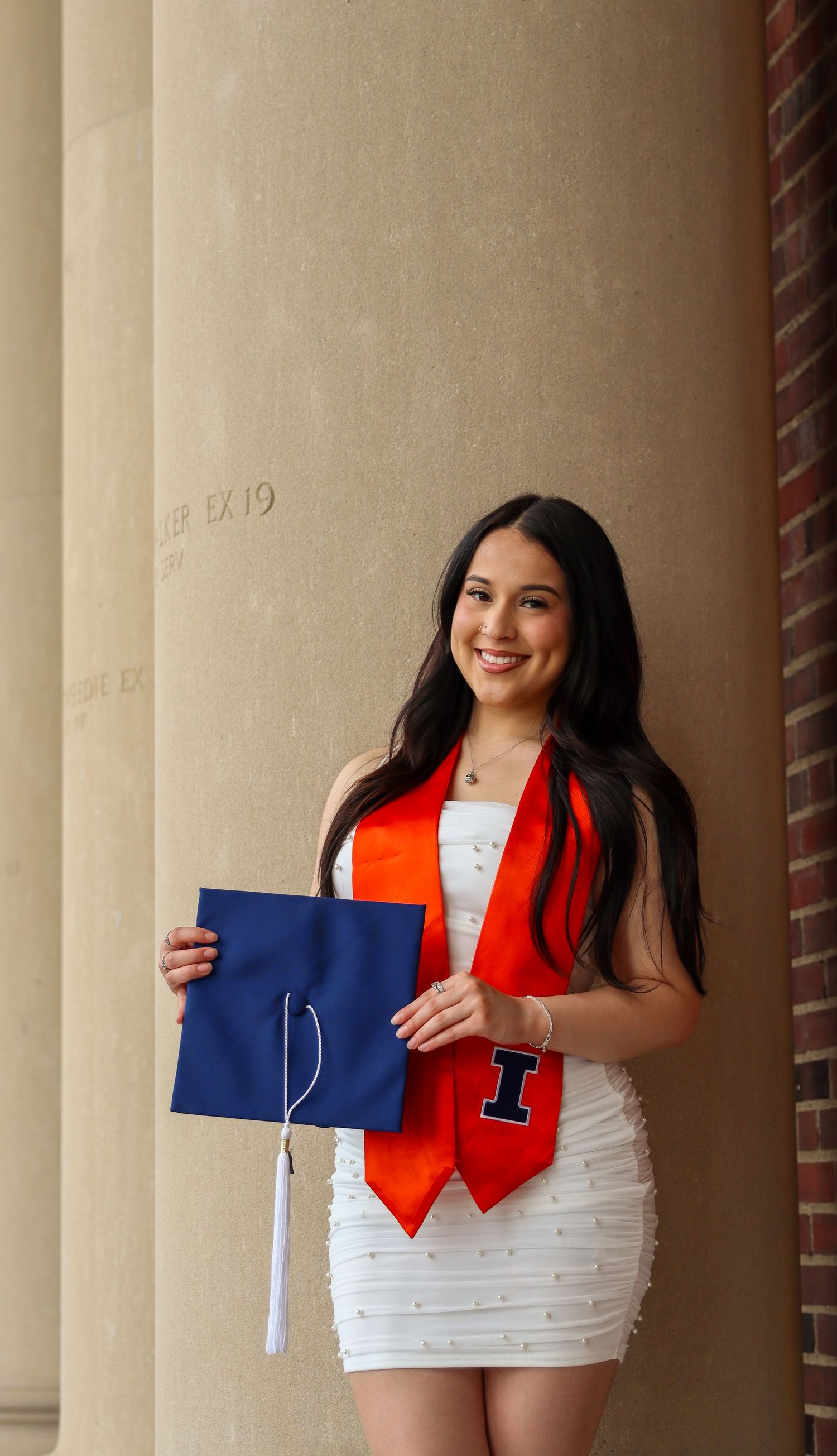 Graduate in white dress and red graduation stole holds blue diploma cover while standing by column.