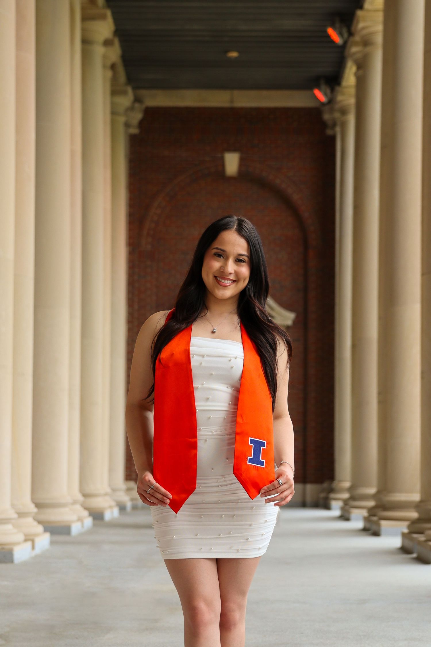 Graduate wearing white dress and orange stole stands in columned hallway during commencement photoshoot.