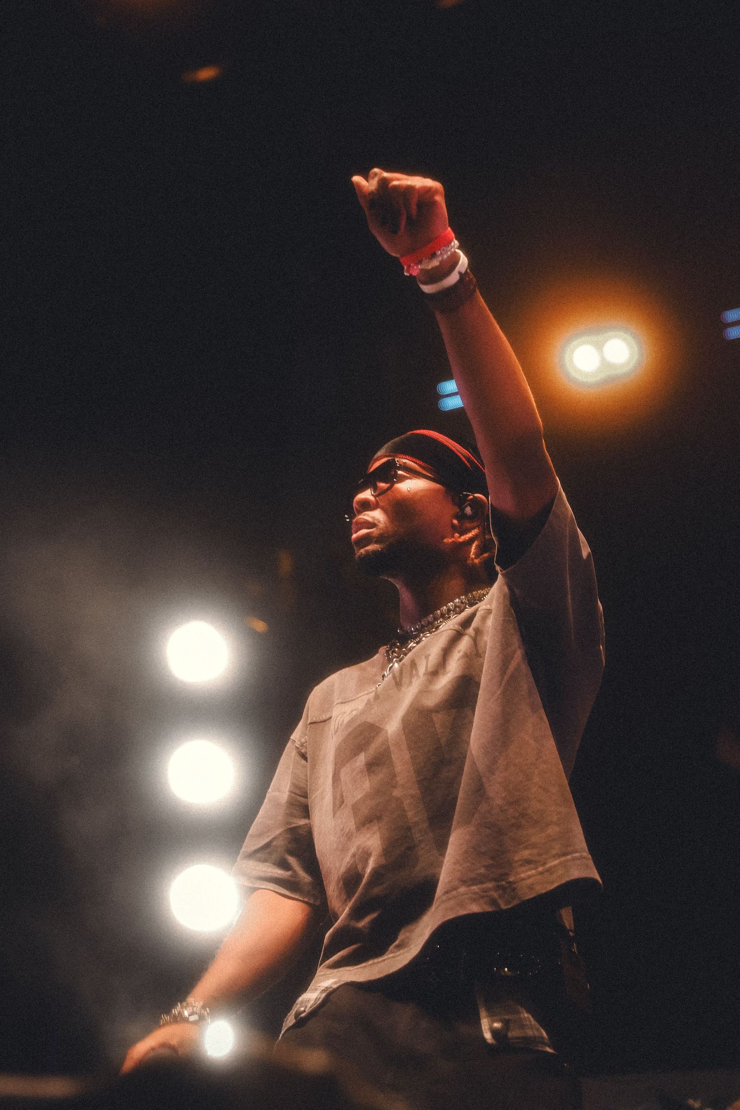 Performer on stage raises fist during concert with dramatic lighting and spotlights in dark atmosphere.