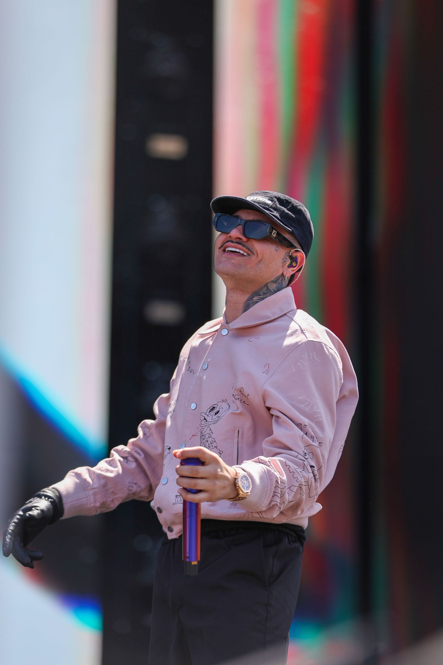 A performer in a pink jacket and baseball cap gestures expressively during an outdoor performance.