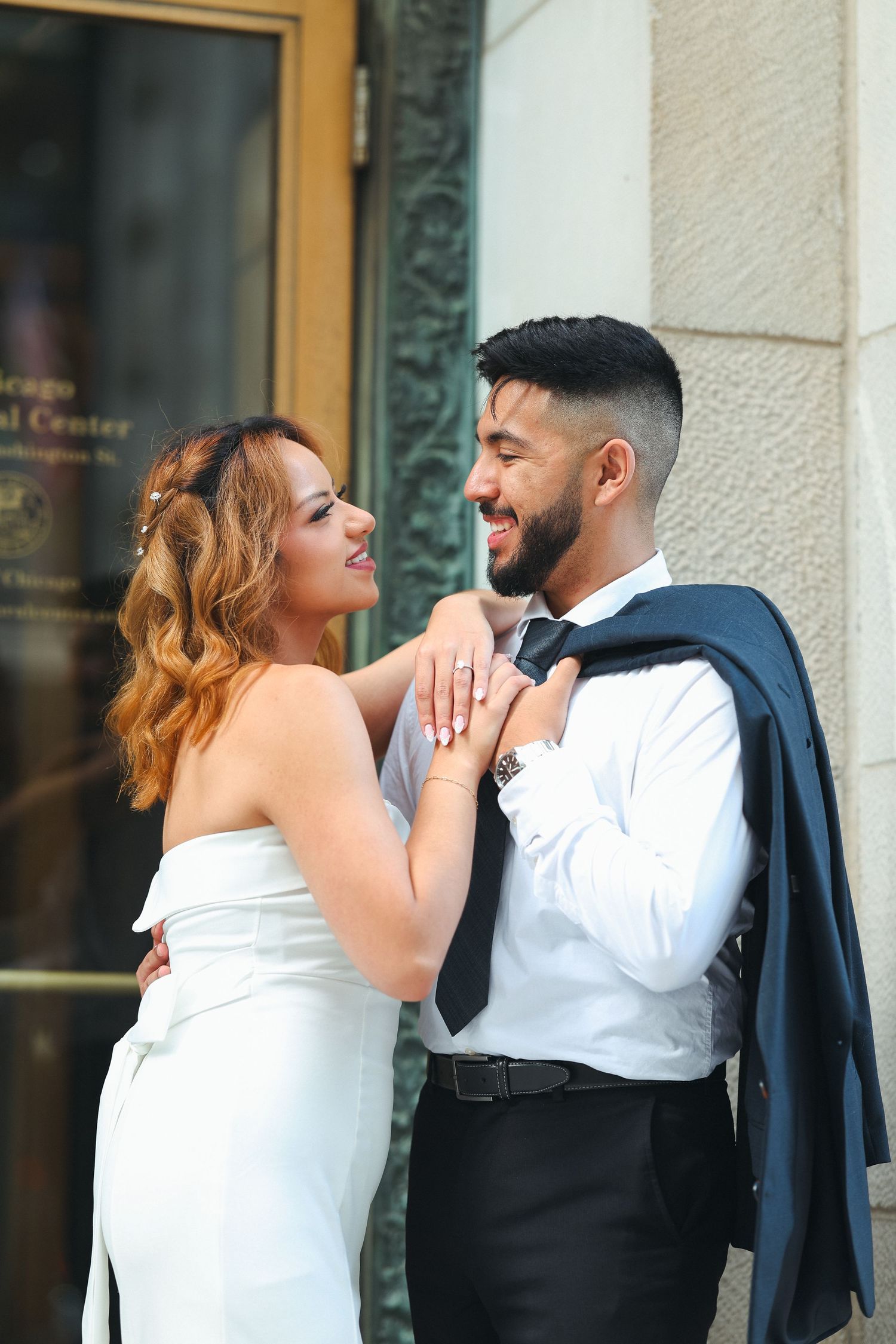 A couple in formal wedding attire share a romantic moment against a storefront wall.