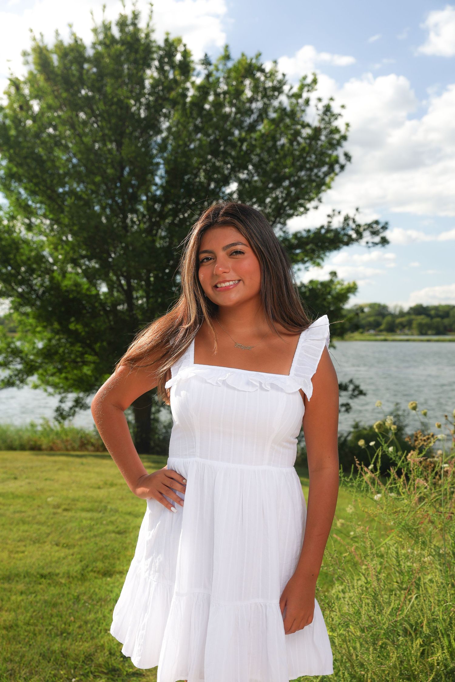 Woman in flowing white summer dress poses outdoors near a lake on a sunny day with green trees in background.