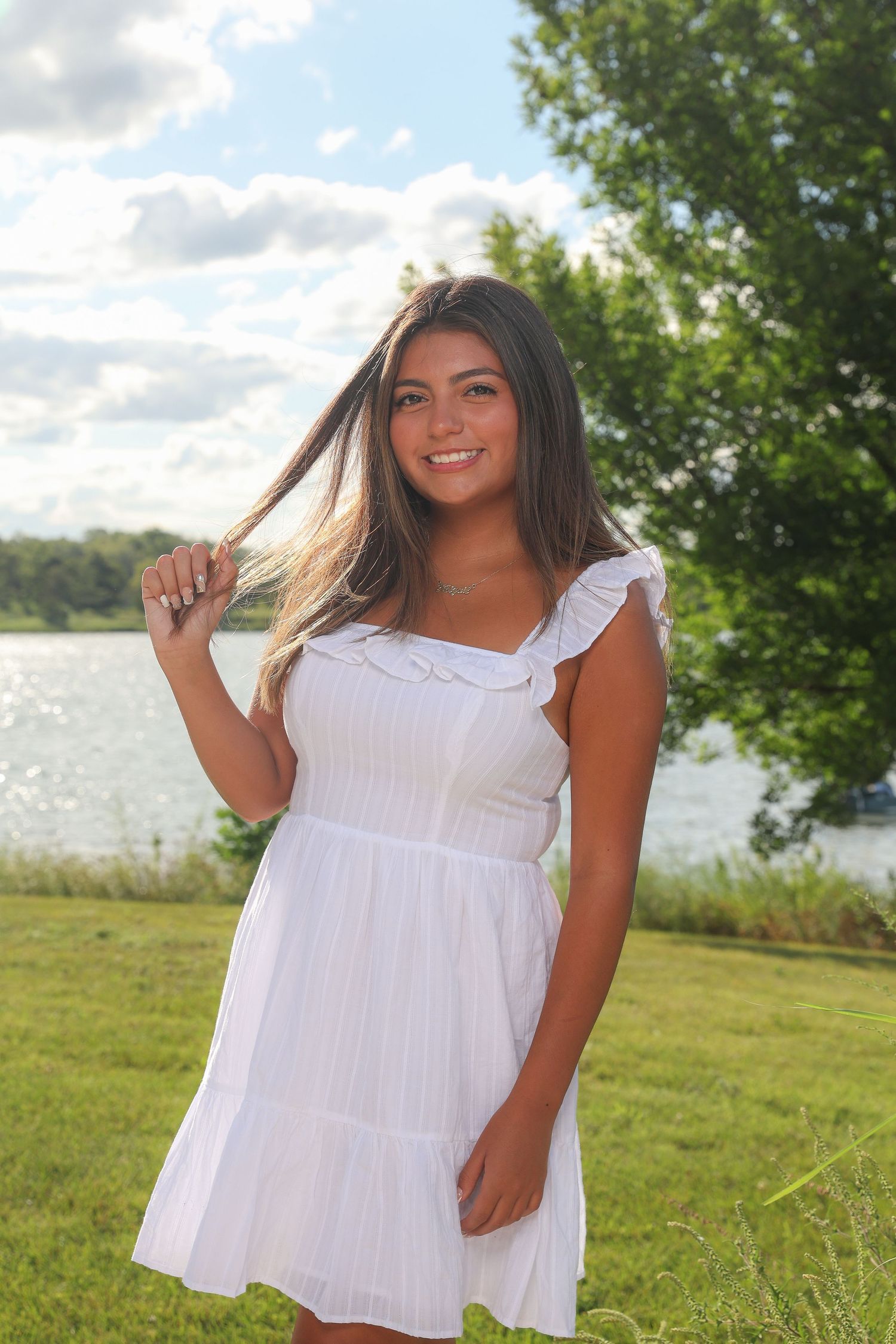 A person in a white summer dress poses outdoors on a grassy hill near a lake on a sunny day.