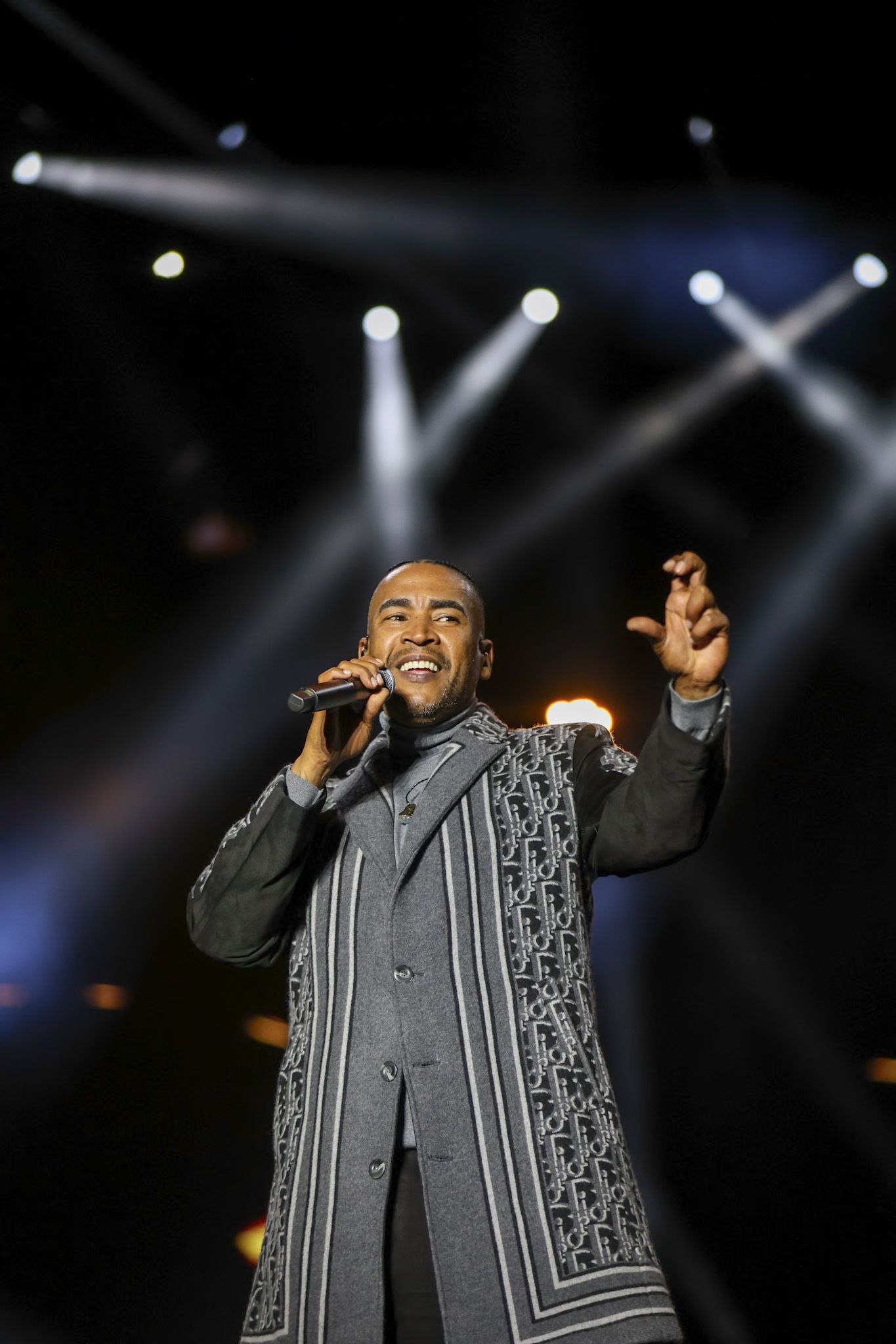 A performer in a gray suit with decorative trim sings into a microphone on stage with dramatic concert lighting beams.
