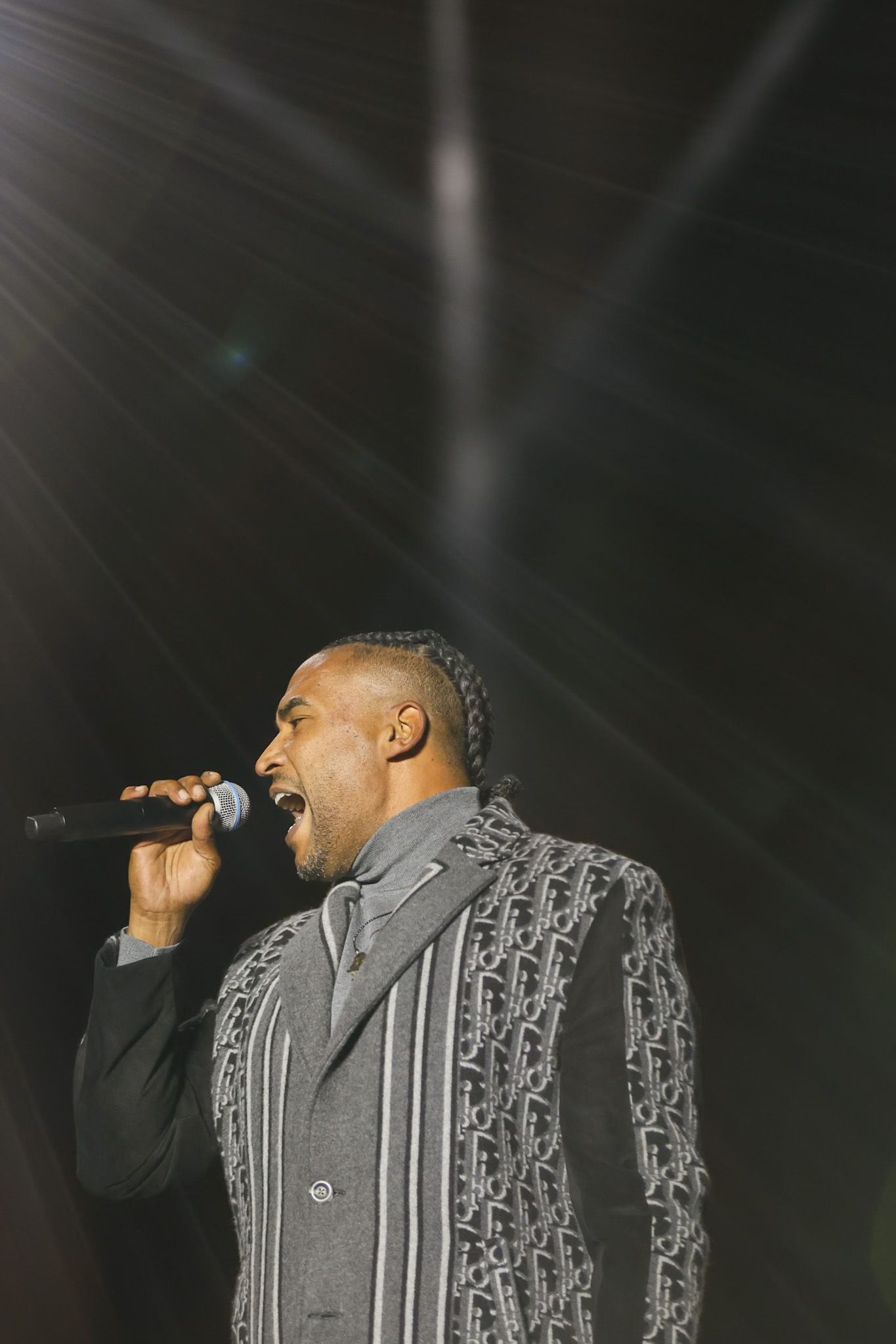 A performer in a gray patterned jacket sings passionately on stage during a concert performance.