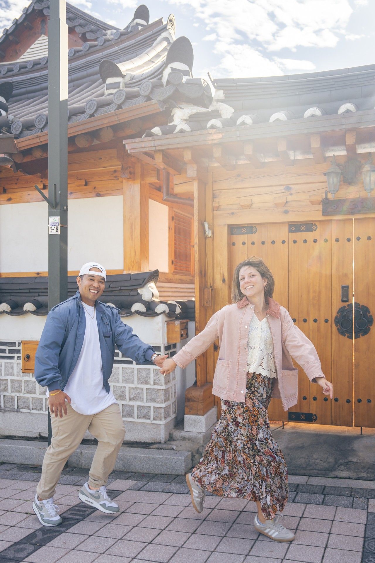Two people in coordinated outfits perform playful dance moves together in front of a traditional Korean building.