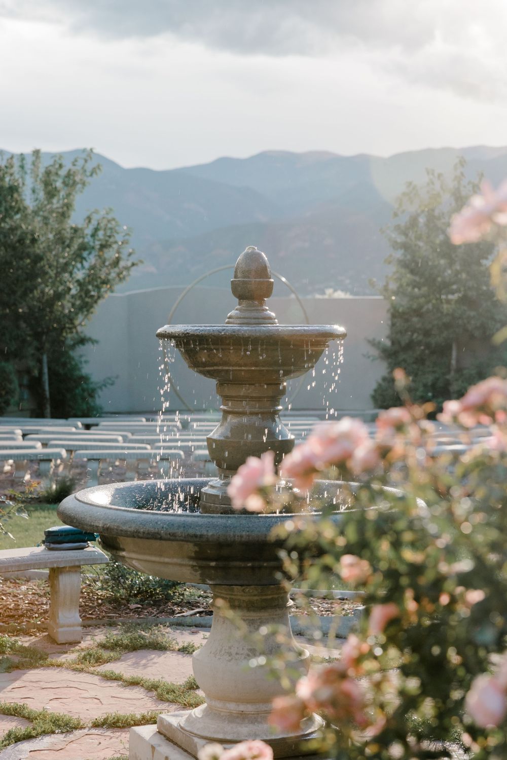 Ornate stone fountain with water jets spraying in a garden with pink flowers and mountains in the background.