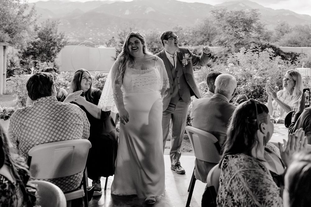 Black and white wedding ceremony photo of a couple walking down the aisle outdoors with guests seated on both sides.