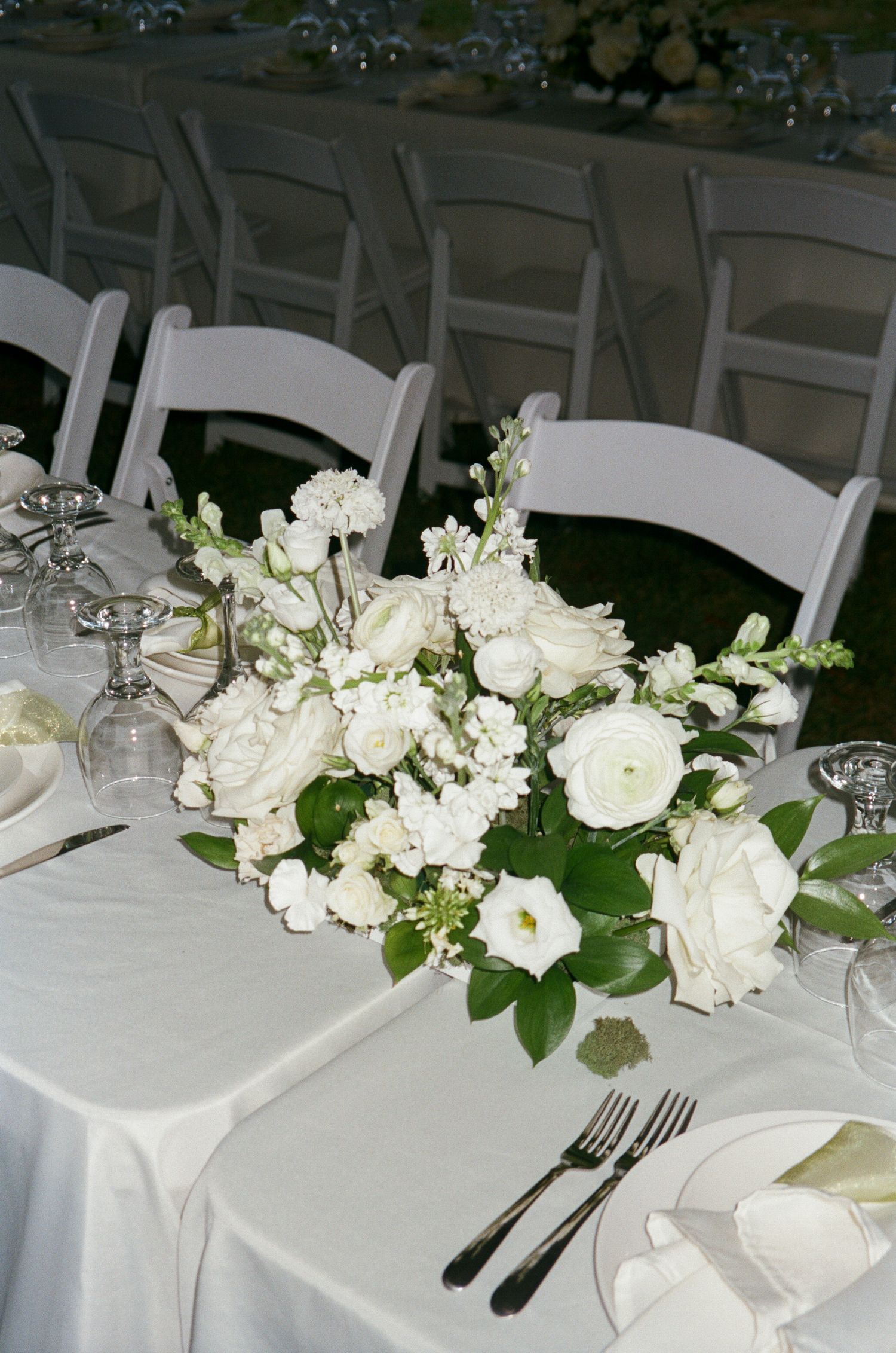 Elegant white wedding table settings with roses and greenery centerpieces on crisp white linens.