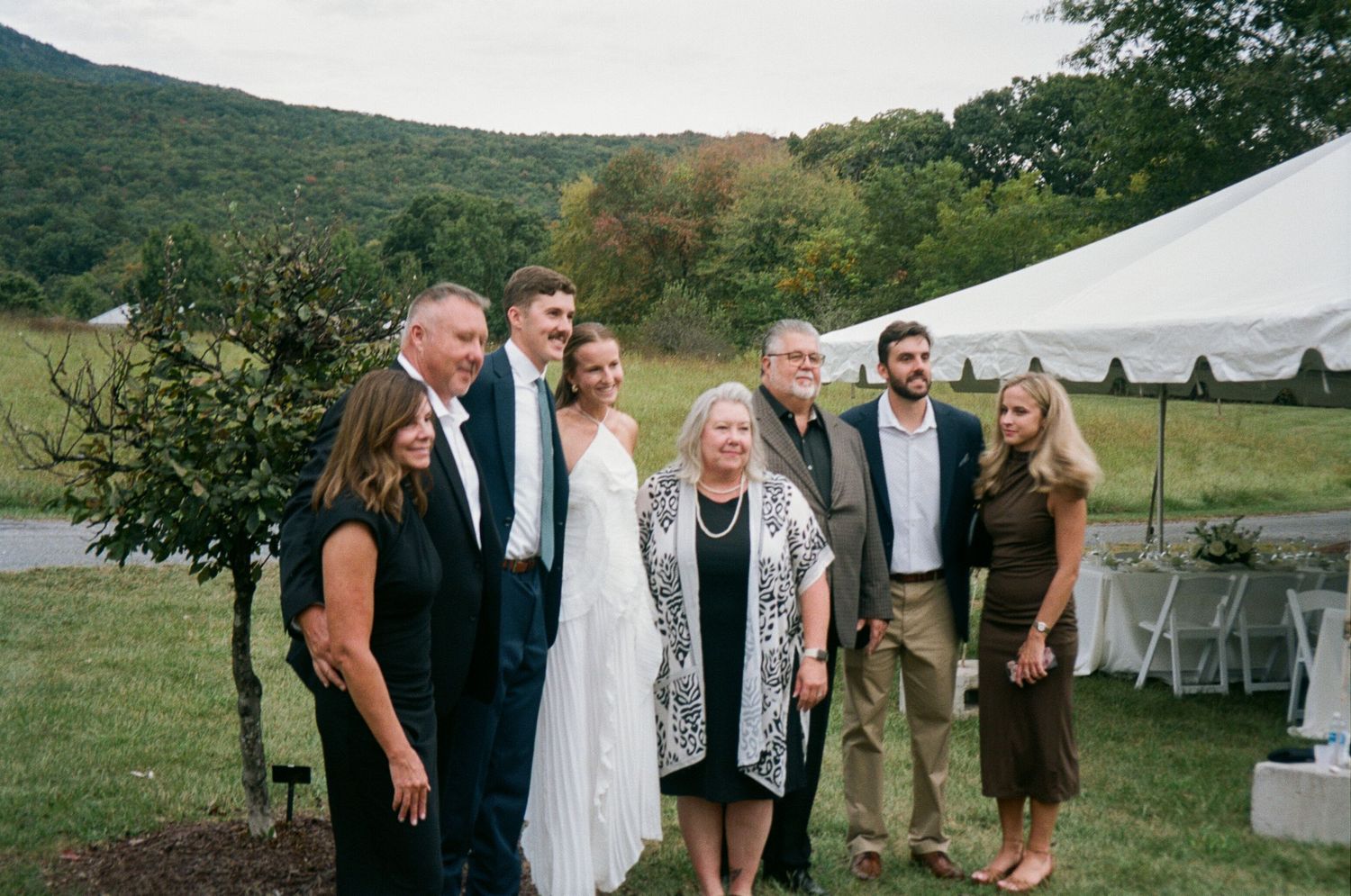 A group of people celebrating outdoors at a wedding reception with a white tent and green hills in the background.