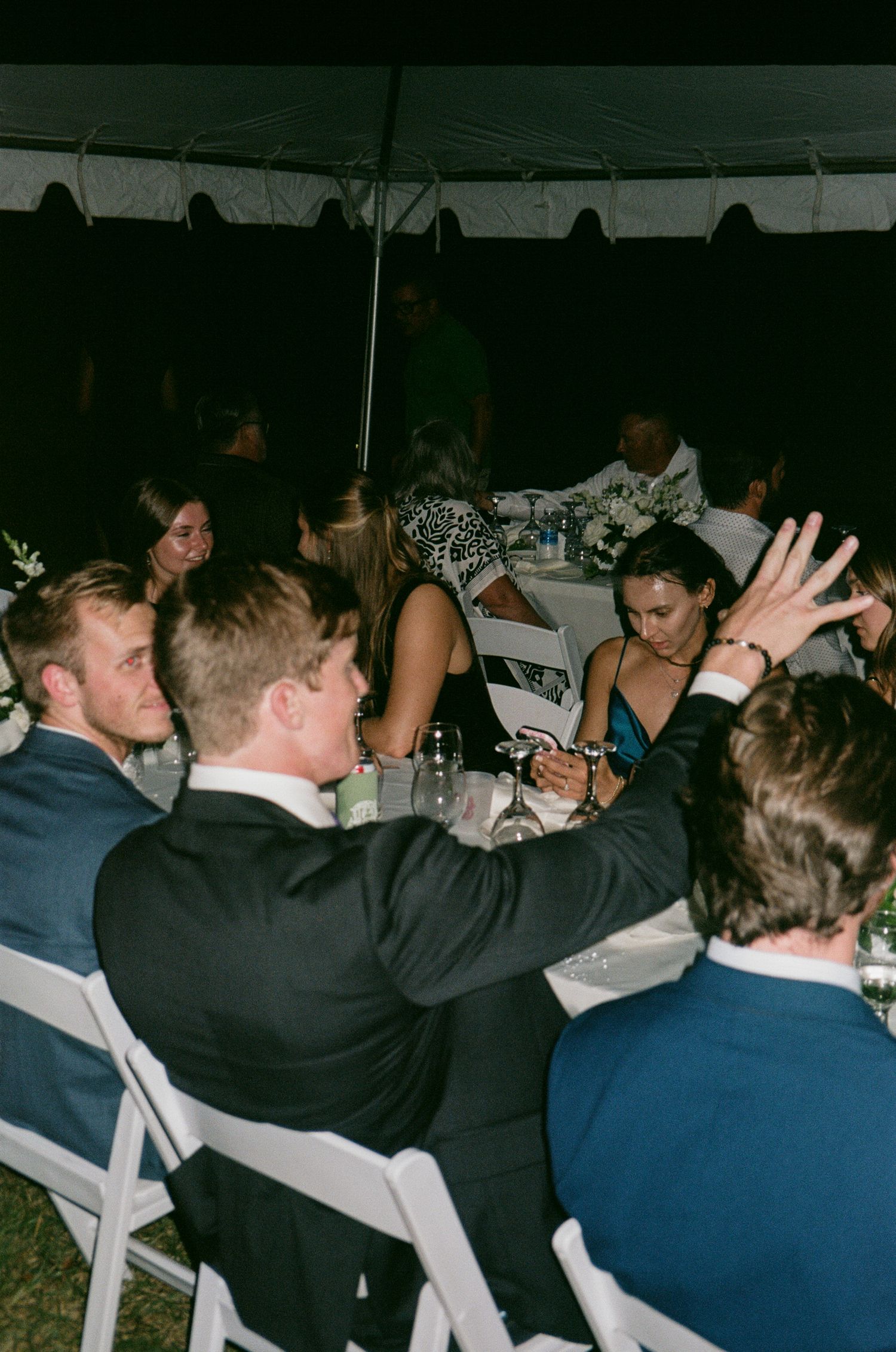 A group of wedding guests seated at tables under a tent raise their hands during a reception celebration.