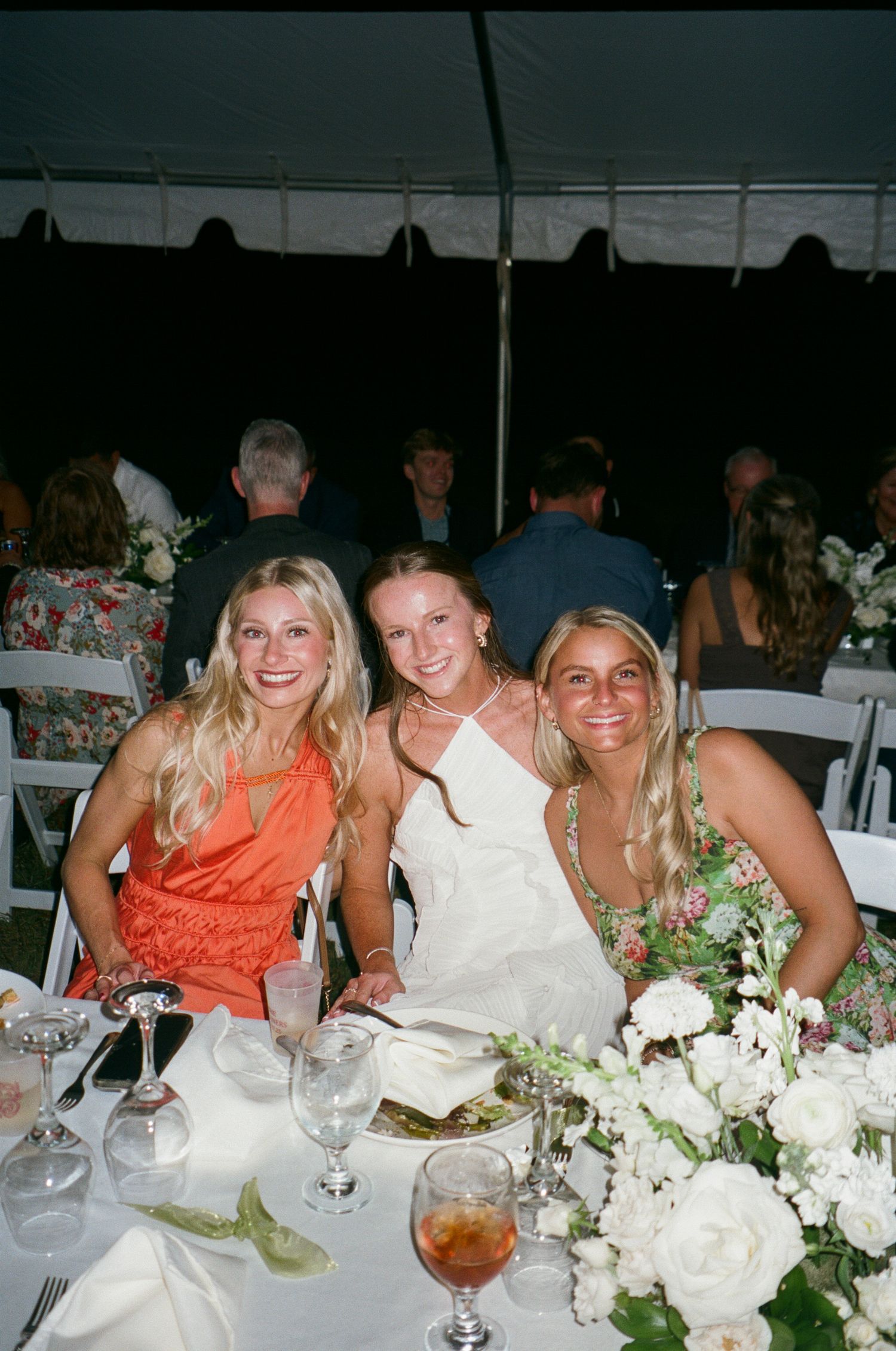 Wedding guests enjoy dinner at an elegantly decorated reception table with white floral centerpieces and glassware.